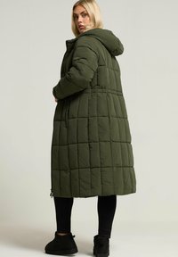 Lång olivgrön puffjacka med huva, med en quiltad design och sidofickor med dragkedja, bärs över svarta leggings och ankelstövlar.