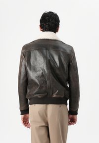 Hnedá kožená bomber bunda s krémovým ovčím golierom, rebrovanými manžetami a lemom. Má mierne lesklú textúru a priliehavý strih.