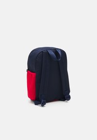 Zaino in tessuto blu navy con una tasca laterale rossa. Presenta spallacci regolabili e una maniglia superiore. Texture liscia; forma rettangolare con bordi arrotondati.