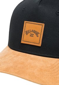Black cap with a tan suede brim, featuring a rectangular leather patch labeled "BILLABONG" in dark lettering. Textured fabric and stitching visible.