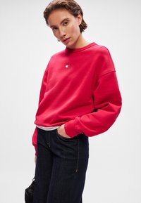 Sweatshirt rouge à col rond et épaules tombantes, avec le logo "TOMMY" au centre. Assorti à un jeans en denim foncé.