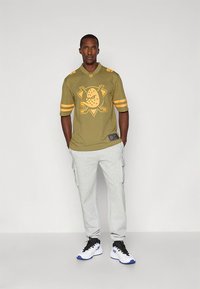 Olivzaļš džersijs ar dzeltenām svītrām un grafisku logo centrā; kopā ar gaiši pelēkām kravas stila sporta biksēm un melniem sporta apaviem.