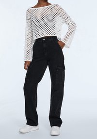 Una modella indossa un top corto bianco a maglia aperta e pantaloni cargo neri a vita alta con tasche, completati da sneakers bianche, su uno sfondo chiaro.