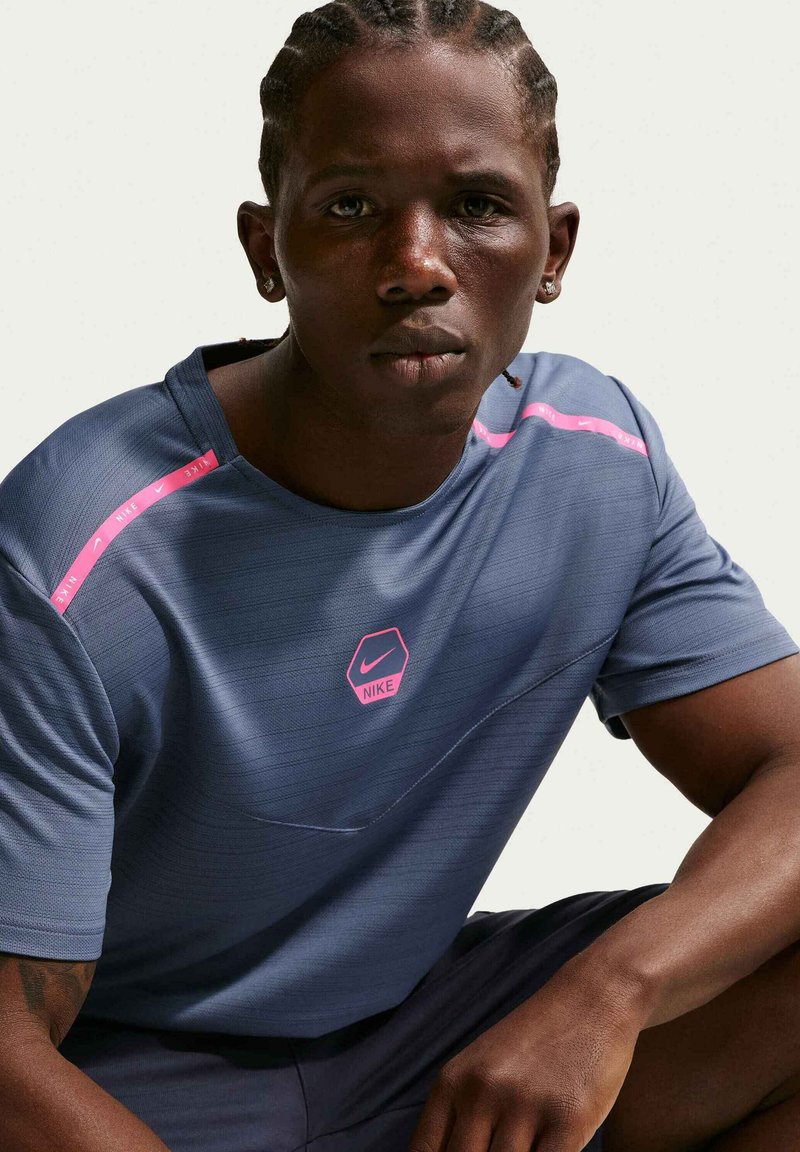 Joven con el cabello trenzado que lleva una camiseta azul de Nike con detalles rosas, sentado y mirando directamente hacia adelante con un fondo liso.