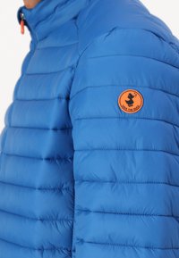 Blauwe pufferjack met horizontale stiksel, voorzien van een oranje rond logo op de linker schouder. Glad, glanzend materiaal met een hoge kraag.