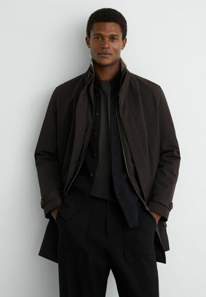 Ung mand iført mørkt lag-på-lag tøj med hænderne i lommerne, stående foran en ensfarvet lys væg, vendt fremad.