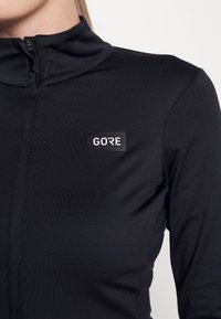 Black athletic long-sleeve top with a high collar and zipper. Features a rectangular logo patch with "GORE" in white lettering. Smooth texture.