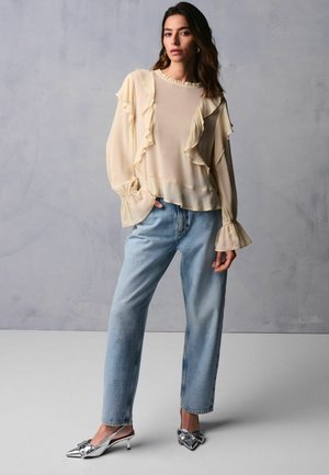 Helle beigefarbene Chiffonbluse mit Rüschenkanten und weit ausgestellten Ärmeln, kombiniert mit blauen, ausgefransten Jeans aus Denim mit silbernen Verzierungen und metallischen Schuhen.