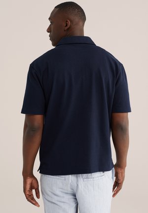 Mężczyzna stojący tyłem, ubrany w granatową koszulkę polo z krótkim rękawem i jasne spodnie, na tle jednolitego beżowego tła.