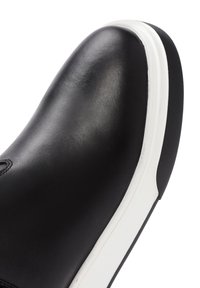 Zapatilla de cuero negra con una textura suave, punta redondeada y suela de goma blanca. Presenta detalles cosidos y un diseño minimalista.