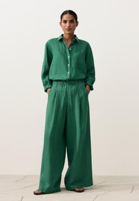 Nő áll zsebre tett kézzel, laza zöld gombos ingben és széles szárú nadrágban, fekete szandálban, semleges háttér előtt.