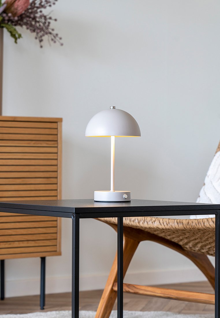 Lámpara de mesa blanca en forma de cúpula iluminada sobre una mesa de metal negro, junto a una silla de madera trenzada y un armario de madera con listones horizontales.