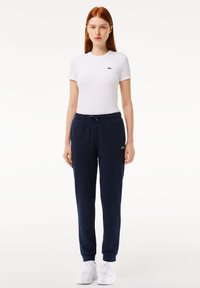 Hvit tettsittende t-skjorte med en liten grønn logo, kombinert med marineblå joggebukser med elastisk midje og mansjetter. Hvite joggesko fullfører antrekket.