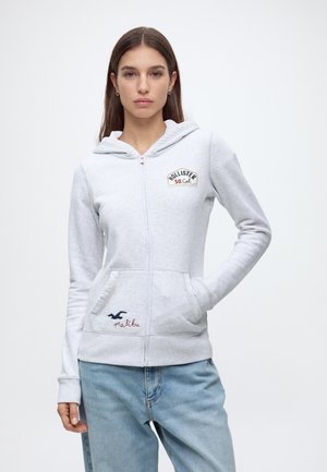 Jeune femme portant un sweat à capuche gris clair zippé avec le logo Hollister So Cal et un jean bleu, debout, la main dans la poche.