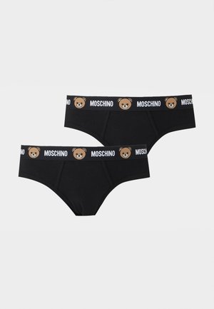 Särgita mees paljajalu, kandes musti pükse, millel on karumõmmiku näod ja „Moschino“ logo vöökohal, ühevärvilise tausta ees.