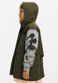 Olivgrön jacka med huva, med en grå ärm som har en svart Mickey Mouse-grafik. Designen inkluderar tryckknappar och sömdetaljer.