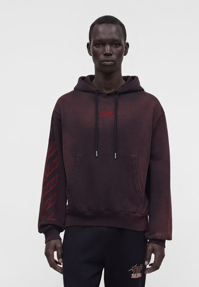 Homme portant un sweat à capuche bordeaux foncé avec des motifs géométriques rouges sur la manche gauche et un petit texte rouge sur la poitrine.