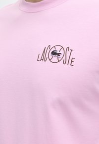 Ροζ ανοιχτό πουκάμισο με το λογότυπο "LACOSTE" και τον κεντημένο μαύρο κροκόδειλο μέσα σε κύκλο στο στήθος.