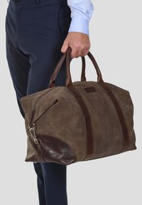 Sac de voyage en suède marron avec des accents en cuir et des poignées supérieures. Il dispose d'une fermeture éclair et d'un détail de couture en diagonale.
