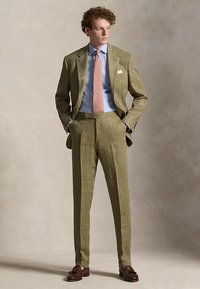 Mann in olivgrüner Anzug, hellblauem Hemd, rosa gemusterter Krawatte und braunen Loafers, steht mit den Händen in den Taschen und schaut nach rechts.