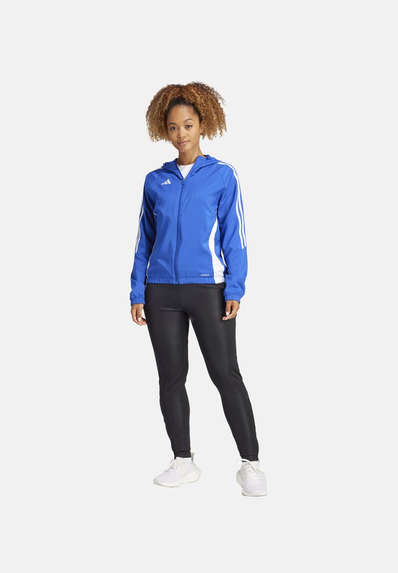 Blauwe sportjack met witte zijaccenten, ritsopening en kap, gecombineerd met zwarte leggings en witte sneakers.