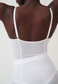Witte bodysuit met verstelbare bandjes en gouden hardware. Kenmerkt zich door een zachte textuur en naadloos ontwerp, dat de silhouet accentueert.