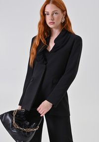 Blazer nero con colletto drappeggiato, due tasche frontali e vestibilità sartoriale. In mano una borsa nera con manico a catena. Texture liscia.