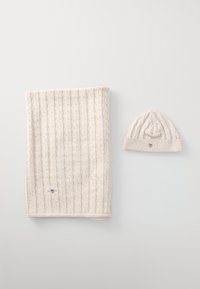 Écharpe en tricot beige avec un motif en relief à côtes, accompagnée d'un bonnet assorti arborant un petit logo et un design à côtes similaire.