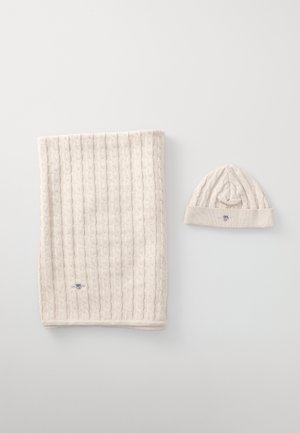 Écharpe en tricot beige avec un motif en relief à côtes, accompagnée d'un bonnet assorti arborant un petit logo et un design à côtes similaire.