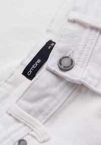 Tejido de mezclilla blanca con un botón metálico con el grabado "Ombre Denim" y una etiqueta que dice "Ombre" y talla "M 32."