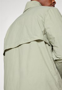 Light green jacket featuring a classic collar, textured fabric, and a layered back design, with a relaxed fit and smooth finish.