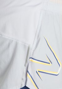 Ljust grå idrottsshorts med en mjuk textur, med blå och gula diagonala ränder och reflexdetaljer på sidan.