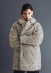 Bontjas in lichtbeige met een hoge kraag, lange mouwen en een losse pasvorm. De textuur is zacht en pluizig, met verborgen zijzakken.
