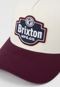 Béžová bé baseballová šiltovka s vínovým šiltom, zdobená vyšitým nášivkom námorníckej modrej farby s textom "Brixton MFG. CO." a detailom v kruhovej forme.