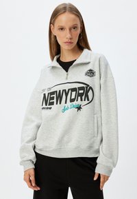 Grå sweatshirt med halvdragkedja, med svarta och teal grafik, framficka samt ribbade muddar och fåll. Mjuk material och avslappnad passform.