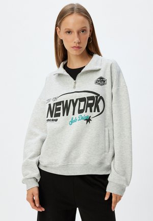 Sudadera gris con cuello medio zip, con gráficos en negro y verde azulado, bolsillo frontal y puños y dobladillo de canalé. Material suave y corte relajado.