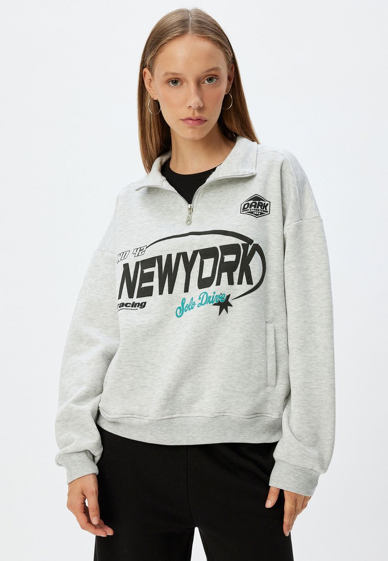 Grå sweatshirt med halvdragkedja, med svarta och teal grafik, framficka samt ribbade muddar och fåll. Mjuk material och avslappnad passform.