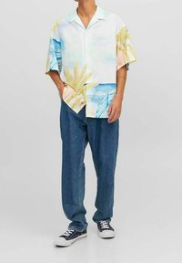 Camicia a maniche corte con bottoni, caratterizzata da una stampa di foglie di palma multicolori su sfondo bianco. Abbinata a jeans blu larghi e sneakers.