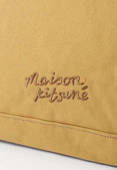 Geantă din pânză galbenă, cu logo-ul brodat "Maison Kitsuné" în nuanțe de brun. Suprafață texturată cu cusături vizibile, care se aliniază cu designul.