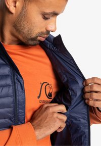 Chaleco acolchado ligero azul marino con una superficie texturizada, llevado sobre una camiseta de manga larga naranja que presenta un logo de ola negra.