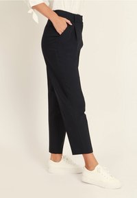 En person klädd i svarta smala byxor med sidofickor, en vit topp med knytärmar och vita sneakers, står med sidan mot en neutral bakgrund.