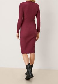 Abito in maglia a coste bordeaux con maniche lunghe, forma aderente, lunghezza al ginocchio e tessuto testurizzato. Indossato con stivaletti neri alla caviglia.
