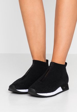 Baskets chaussettes en maille noire avec semelles blanches portées sur des jambes nues sur un fond clair uni.