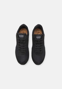 Chaussures de sport noires avec une tige en mesh, une semelle intérieure en liège rembourrée, des lacets noirs et une semelle extérieure en caoutchouc texturée avec un logo sur la languette.