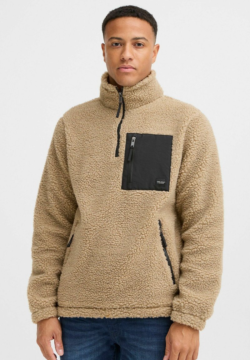 Tan fleece pullover with high collar, black zippered chest pocket, and side pockets; textured, soft fabric with a relaxed fit.