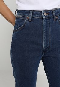 Närbild av en person som bär högmidjade mörkblå denimjeans med kopparnitar och en silverknapp, i kombination med en instoppad vit skjorta.