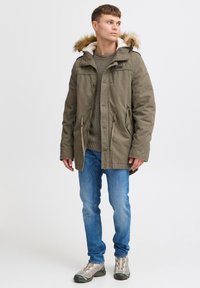 Parka vert olive avec capuche doublée de fourrure, fermeture éclair et bouton, taille élastique, associée à un pull en tricot et un jean bleu clair.
