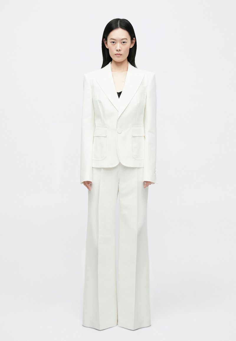 Wit op maat gemaakt pak met een aangesloten blazer met een enkele knoop, opgestikte zakken en broek met wijde pijpen. Glad stof met een matte afwerking.