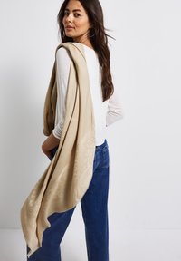 Beige sjaal over de schouder gedrapeerd, zachte textuur met een subtiel decoratief patroon, gecombineerd met een witte lange mouw top en blauwe jeans.