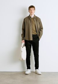 Camicetta grigia scura sopra una maglietta verde, abbinata a pantaloni neri e sneakers bianche. Tiene una borsa a tracolla beige. Design semplice e moderno.